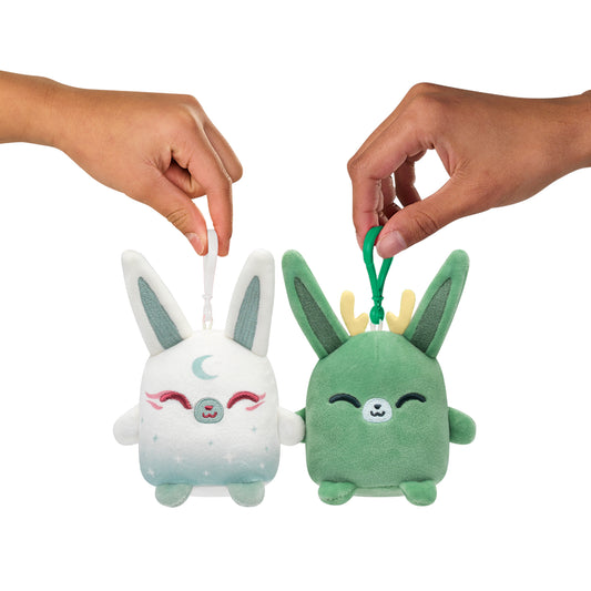 Two hands holding Plushiverse Jackalope & Moon Rabbit Plushmates Besties keychains from the TeeTurtle collection, shaped like cartoon animals with rabbit ears—one light grey, one green—against a white background.