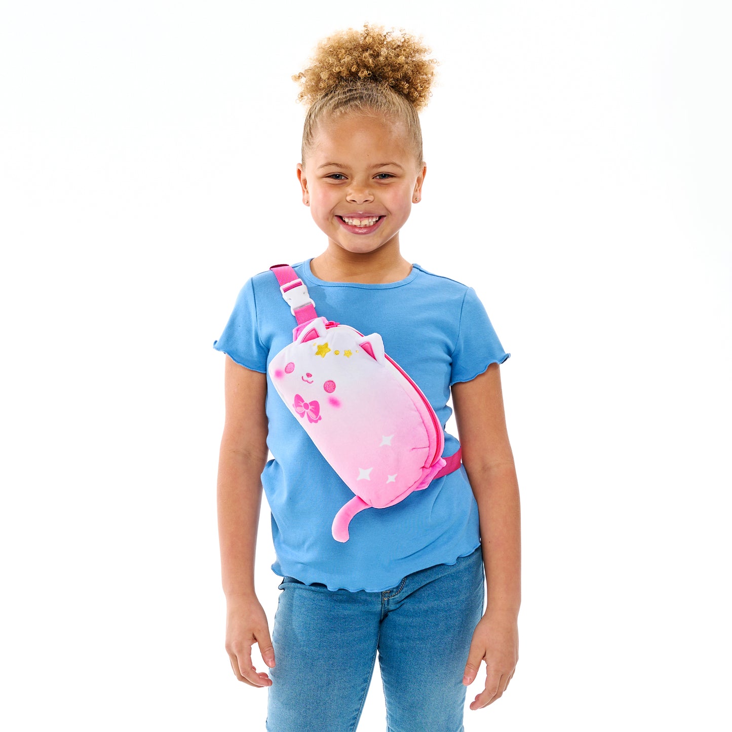 A smiling young girl with a ponytail wearing a blue shirt and jeans and carrying a pink Plushiverse Just Like Meowgic Plushie Fanny Pack from the TeeTurtle collection.