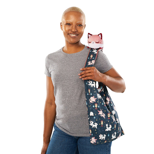A smiling bald woman in a gray t-shirt and jeans, carrying a navy Plushiverse Enchanting Kitsune Tote Bag with cat designs, with a TeeTurtle plushie peeking out.