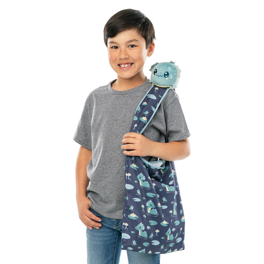 Young boy smiling, holding a blue backpack with Myths & Cryptids collection designs and a Plushiverse Elusive Nessie Plushie Tote Bag on his shoulder, isolated on a white background.
