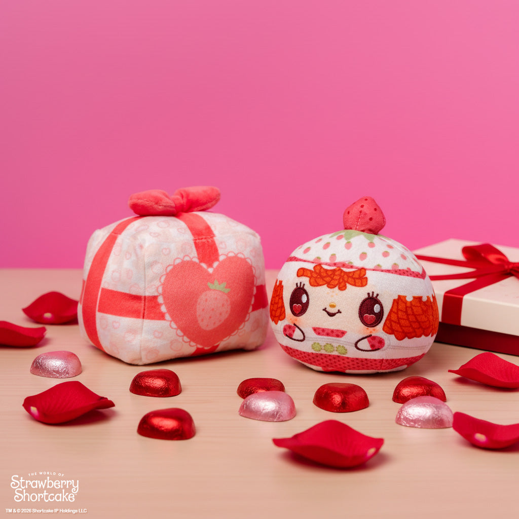 Valentine Present Strawberry Shortcake 4" Strawberry-Scented Reversible Plushie on a table surrounded by red petals, chocolates, and a box with a red bow, with a pink background