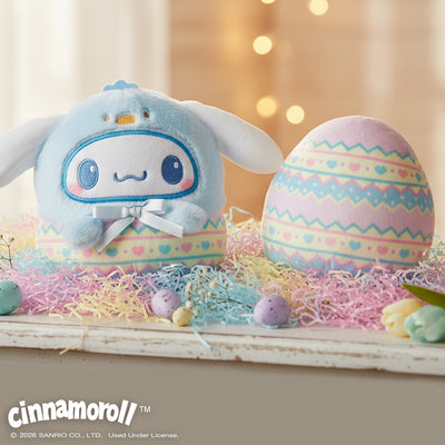 Easter Egg Cinnamoroll 4" Reversible Plushie sitting on a table of pastel paper grass, flowers, and small chocolate eggs with a background of a white curtain and fairy lights