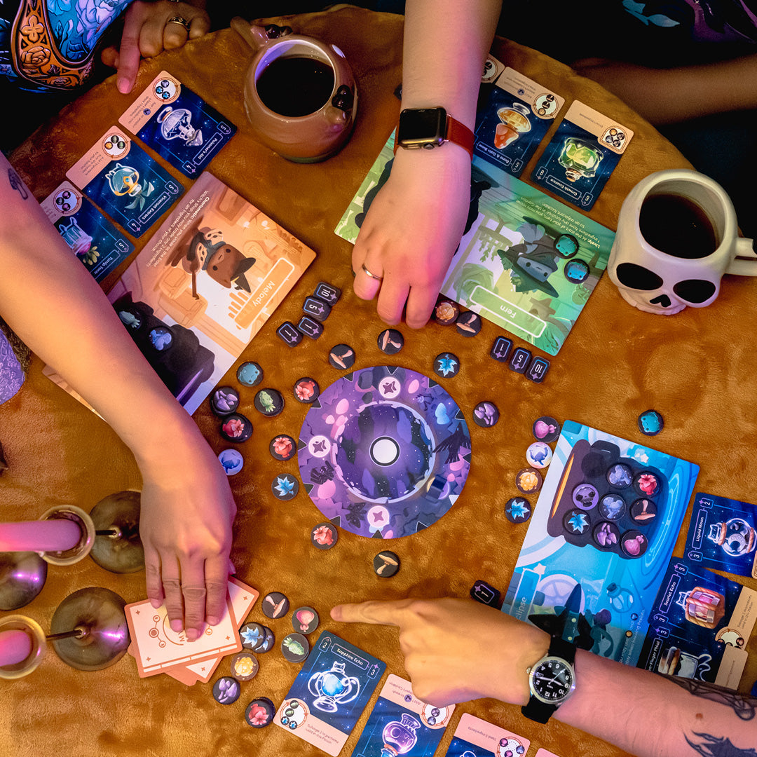 Components of Mystic Moon in action on a tan tablecloth, being played. Only the player's hands are visible. There are also candles and coffee cups on the table in addition to the player board, cards, and tokens. 