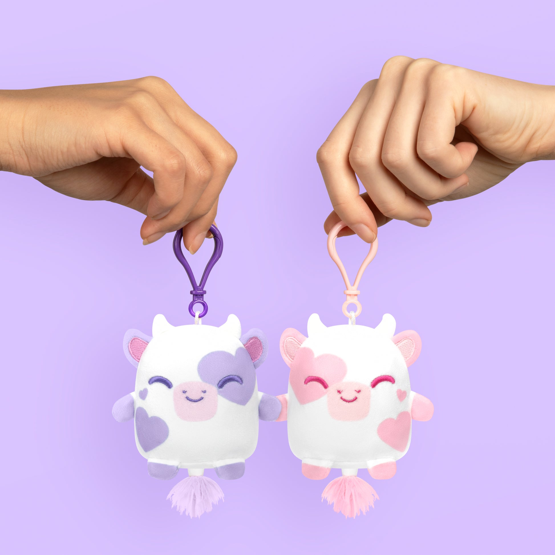 Two magnetic hands holding a Plushiverse I Love Moo Plushmates Besties keychain on a purple background. (TeeTurtle)