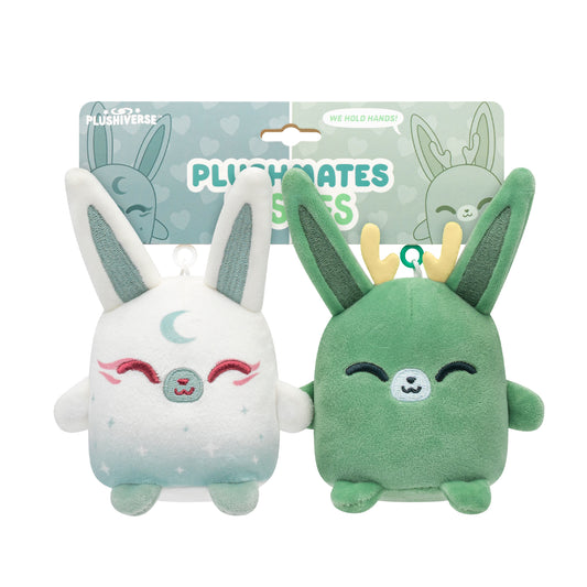 Two Plushiverse Jackalope & Moon Rabbit Plushmates Besties keychains, one white with a moon design and one green with antlers, both with closed eyes and smiling faces, connected by their hands, against a cardboard package. Created by TeeTurtle.
