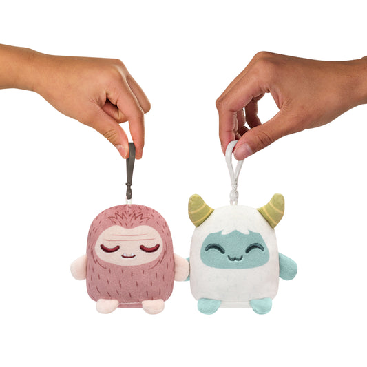 Two hands with magnetic attachments holding Plushiverse Yeti & Bigfoot Plushmates Besties keychains, one pink and one turquoise with horns, against a white background by TeeTurtle.