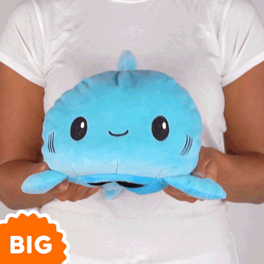 A woman is holding a TeeTurtle Big Reversible Shark Plushie (Blue + Light Blue) among her collection of big mood plushies.