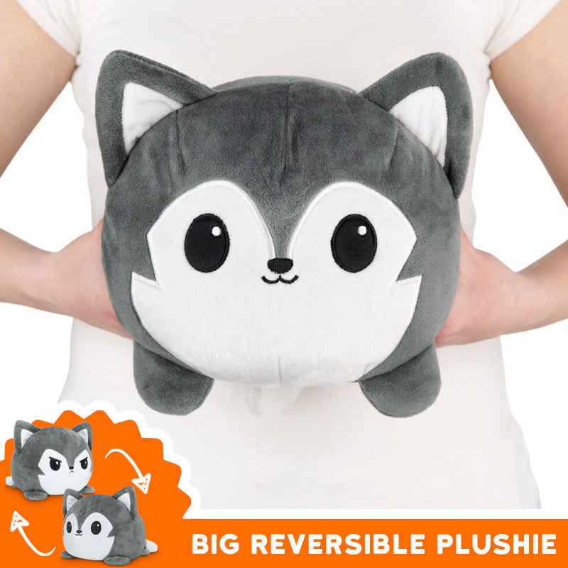 Person holding a big, round, gray, and white plushie with large eyes and fox-like features. Bottom left shows the plushie reversed to reveal different color patterns. Caption reads: "TeeTurtle Big Reversible Wolf Plushie (Gray) by TeeTurtle".
