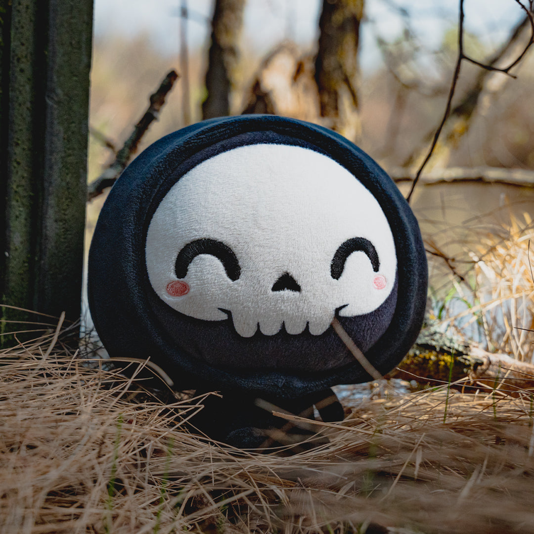 Cloudheads: 5" Grim Reaper (Grim) - A black hooded plush grim reaper with a white skull face, black embroidered eyes and mouth, and small pink cheeks, shown in a wooded area with straw, pine trees and bare trees. 