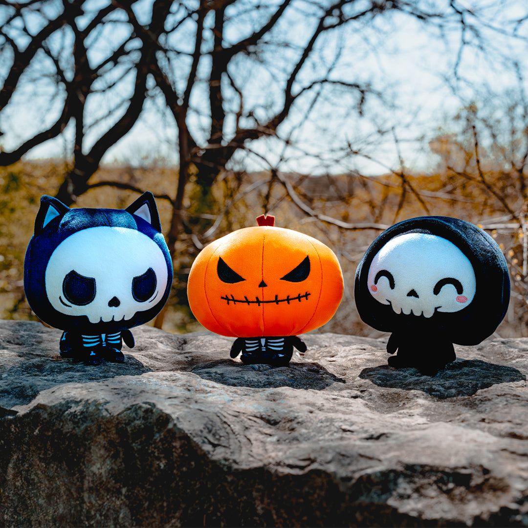 Cloudheads: 5" Halloween Bundle. All three Halloween Cloudheads plushies sitting on a rock in front of bare trees. There is Skelly, the skeleton cat; Punkin, a jack-o-lantern with a skeletal body, and Grim, a cute grim reaper with happy eyes and pink cheeks. 