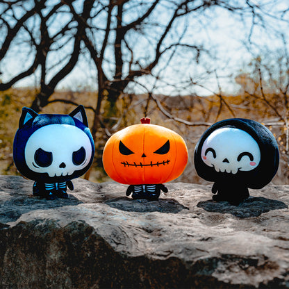 Cloudheads: 5" Halloween Bundle. All three Halloween Cloudheads plushies sitting on a rock in front of bare trees. There is Skelly, the skeleton cat; Punkin, a jack-o-lantern with a skeletal body, and Grim, a cute grim reaper with happy eyes and pink cheeks. 