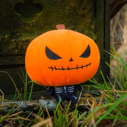 Cloudheads: 5" Pumpkin (Punkin) - A pumpkin headed skeleton plushie with a bright orange head, black stitched eyes and mouth, a small brown stem, and a black-and-white skeleton body. It's outside sitting on a branch with some grass in the foreground and a mossy headstone style stone behind it. 