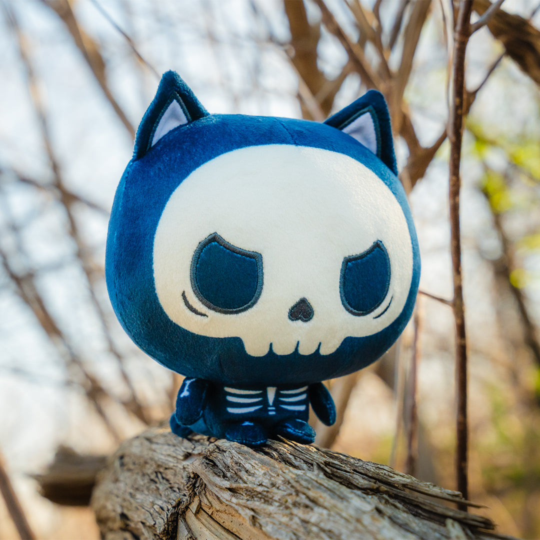 Cloudheads: 5" Skeleton Cat (Skelly) is a cat skeleton Cloudheads plushie. The skull face has a grumpy look to it. It's sitting on a log and there are bare trees in the background. 