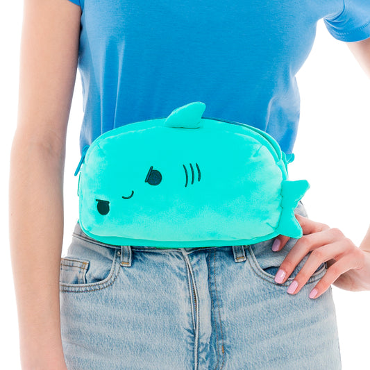 Woman in a blue shirt holding a cute turquoise Plushiverse Jaw-some Shark plushie fanny pack from TeeTurtle against her hip, isolated on a white background.