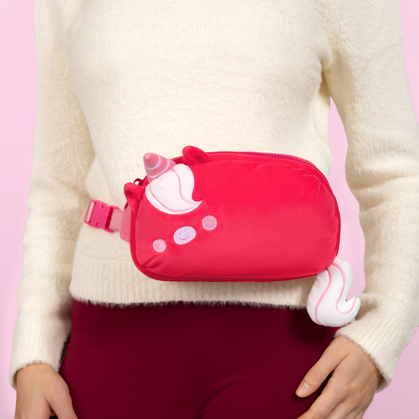 A woman is holding a Plushiverse Ruby Red Unicorn Plushie Fanny Pack by TeeTurtle with hands-free functionality.