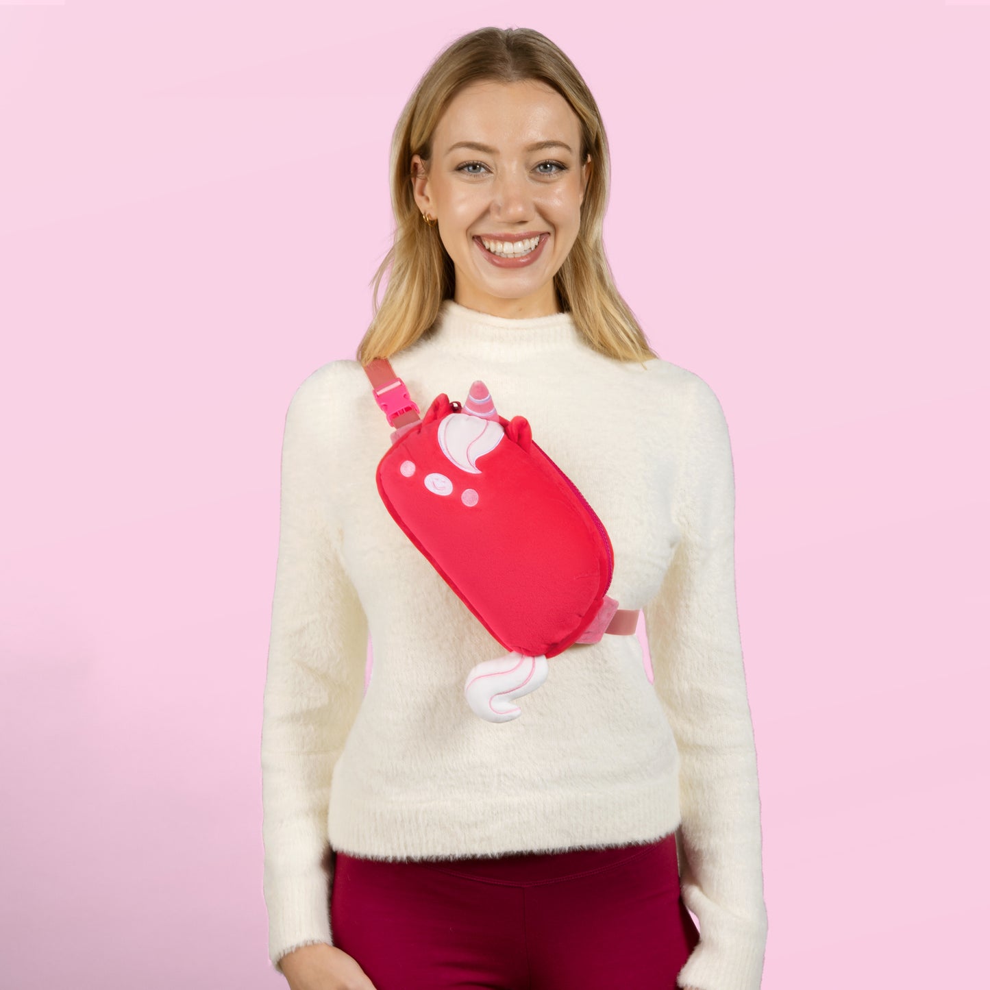 A woman wearing a Plushiverse Ruby Red Unicorn Plushie Fanny Pack by TeeTurtle, featuring a cat design, with adjustable belt for hands-free functionality.