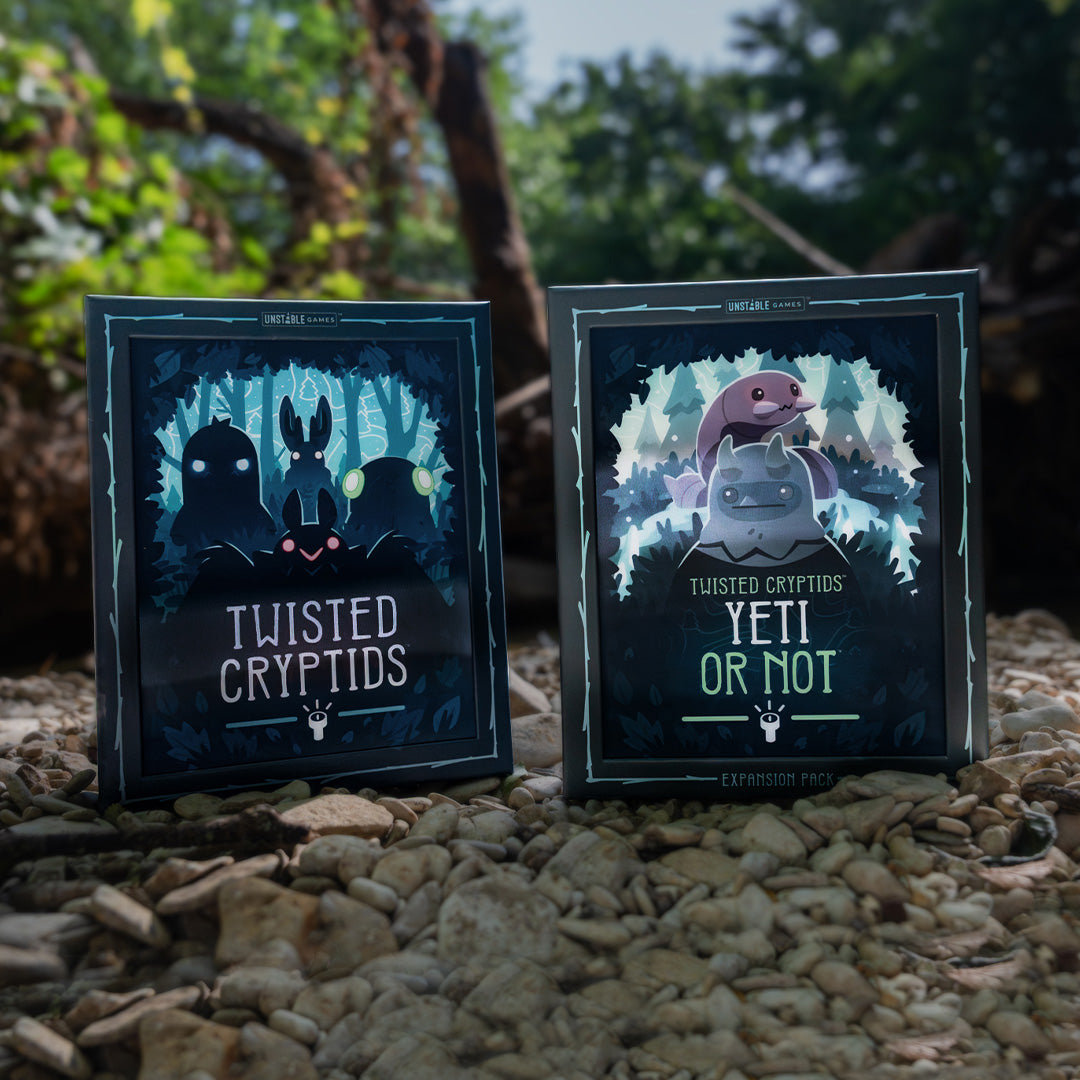 Boxes for Twisted Cryptids and Twisted Cryptids: Yeti or Not expansion stand on a bed of small stones outdoors, with blurred trees and branches in the background. 