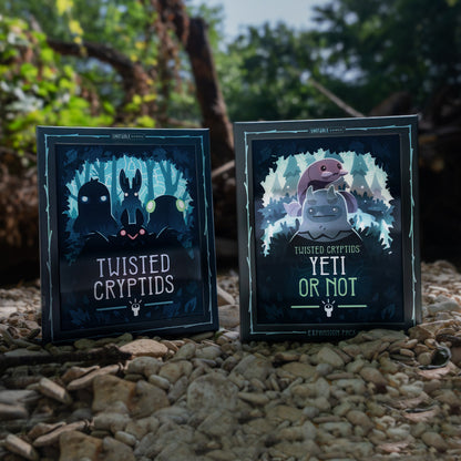 Boxes for Twisted Cryptids and Twisted Cryptids: Yeti or Not expansion stand on a bed of small stones outdoors, with blurred trees and branches in the background. 
