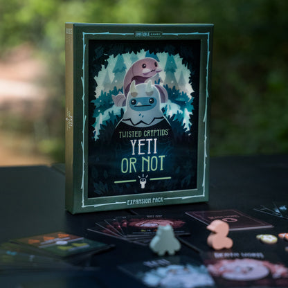 Twisted Cryptids: Yeti or Not Expansion lifestyle image of the box with meeples and cards laid on in front of it and nature behind