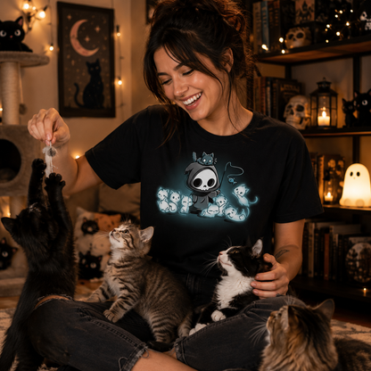 Premium Cotton T-shirt_TeeTurtle Grim and Kitties black t-shirt featuring a hooded skeleton figure, resembling a spooky Grim Reaper, standing surrounded by glowing ghostly cats. One cat is perched on its head while it playfully dangles a toy fish in front of the others.