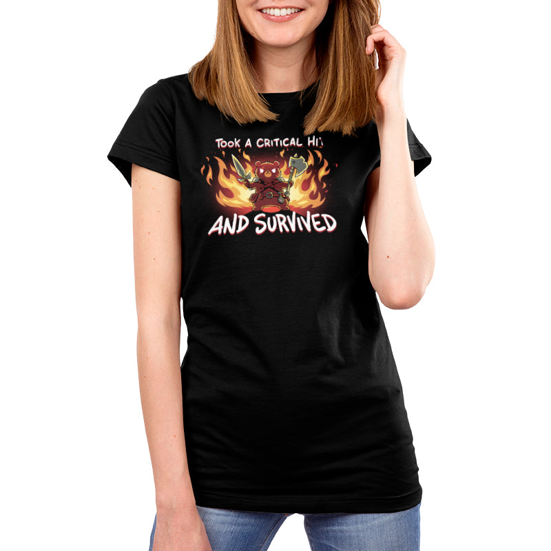 A person wearing a super soft ringspun cotton black T-shirt with the text "TOOK A CRITICAL HIT AND SURVIVED" and an illustration of a character surrounded by flames. The T-shirt is the "I Took A Critical Hit and Survived" by monsterdigital.