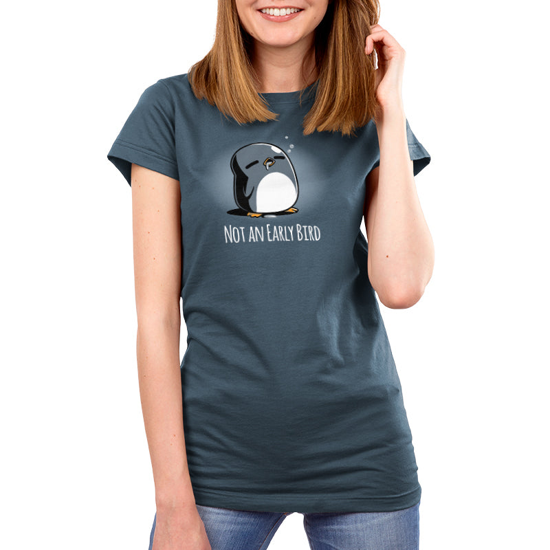 A woman wearing a denim blue, super soft ringspun cotton t-shirt with a sleepy cartoon penguin and the text "Not an Early Bird" stands and smiles. The product she's wearing is the Not an Early Bird by monsterdigital.
