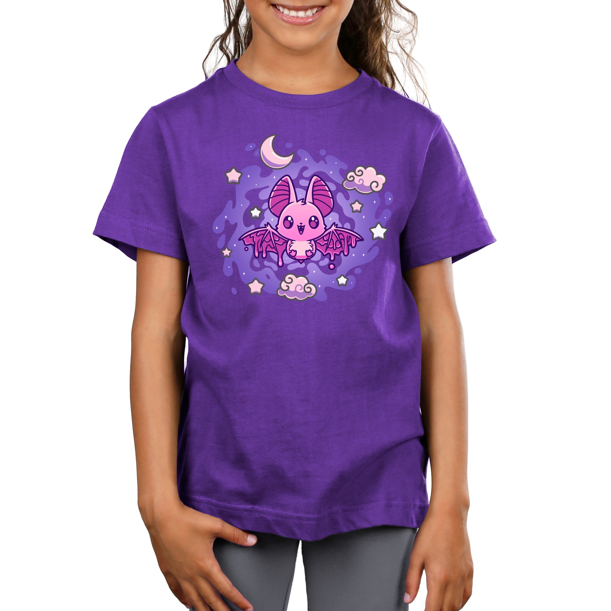 A young child wearing a purple kids T-shirt featuring a pink cartoon bat with stars, clouds, and a crescent moon in the background. The T-shirt is called Itty Bitty Bat and is from the brand monsterdigital.