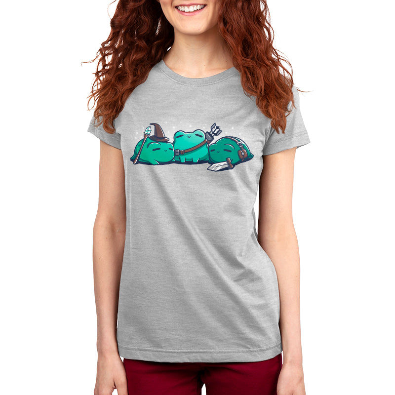 A person wearing a gray kids' T-shirt featuring a cartoon graphic of three green characters, two sleeping and one holding a fork. The individual has long, wavy red hair and is smiling. The T-shirt is the RPG Frogs by monsterdigital.
