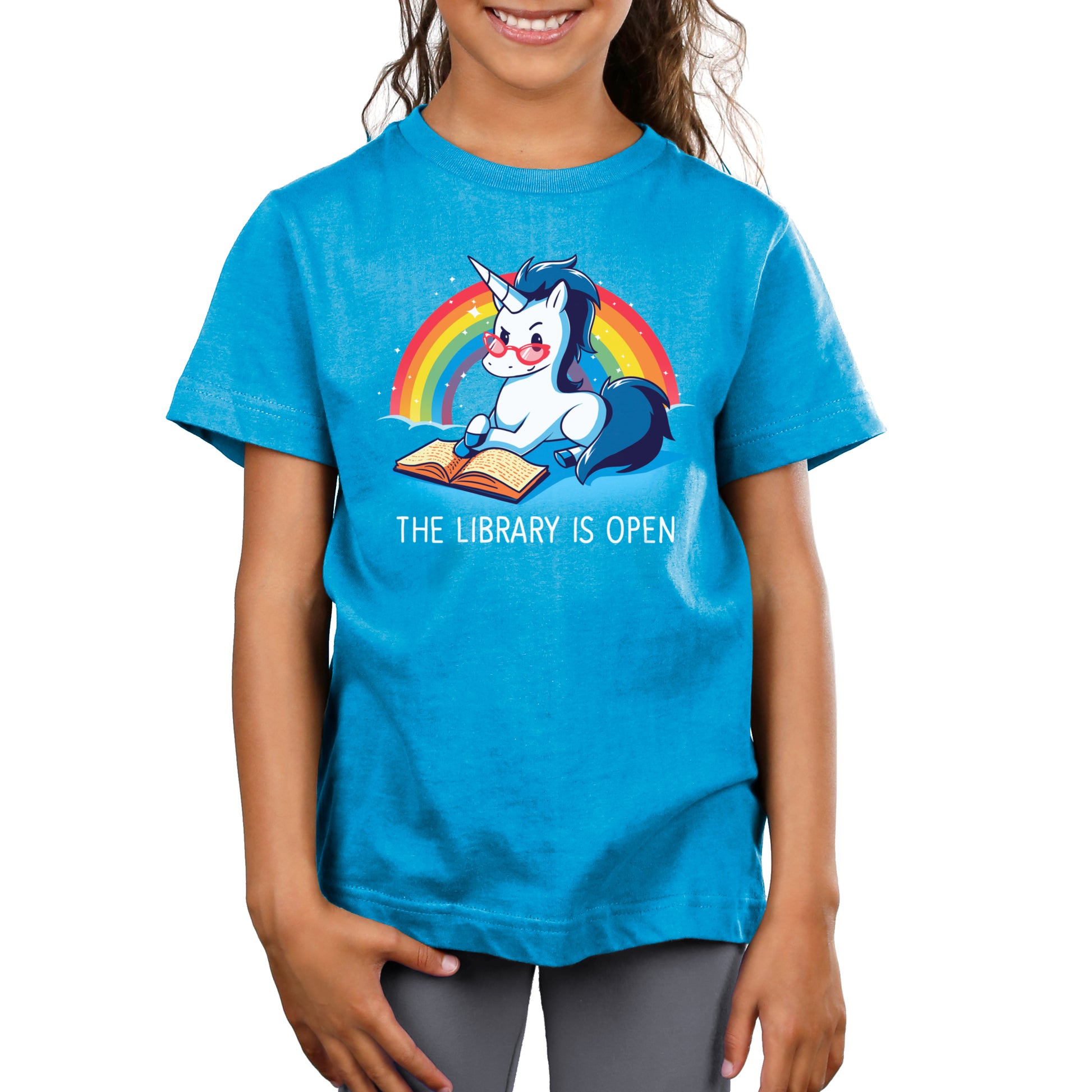 A child wearing a cobalt blue T-shirt with a unicorn reading a book against a rainbow background. Made from super soft ringspun cotton, the shirt proudly displays the text "The Library is Open." This shirt is called The Library is Open by monsterdigital.