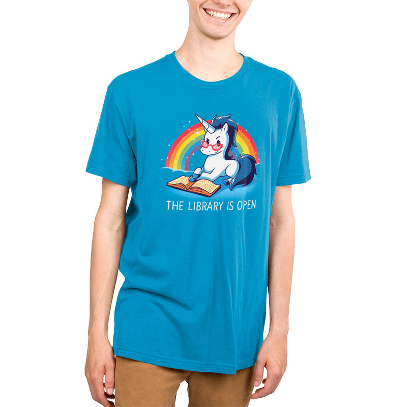 A person wearing a cobalt blue t-shirt with a cartoon unicorn reading a book under a rainbow captioned "The Library Is Open" from monsterdigital. The shirt, called The Library is Open, is made from super soft ringspun cotton for ultimate comfort.