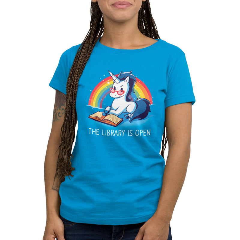 A person wearing a cobalt blue "The Library is Open" t-shirt by monsterdigital, featuring a cartoon unicorn reading a book, with a rainbow in the background. The super soft ringspun cotton makes it as comfortable as it is charming.