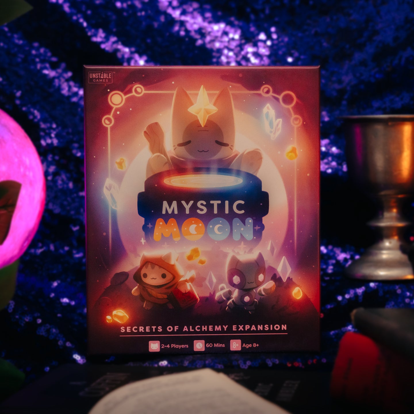 Mystic Moon Alchemy Expansion - the box is tones of yellow, red and purple and has a witch cat above a cauldron with two other witch cat characters below it. It's in front of a glittery purple background and there are spooky props at the edges of the image