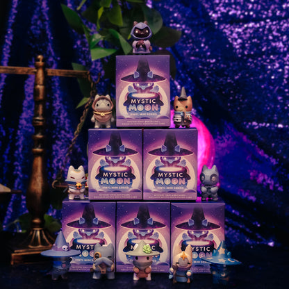 A display of Mystic Moon Vinyl Figure Blind Boxes, arranged in a pyramid with vinyl figures on top of and in front of it. The background is sparkly purple fabric and an antique scale and leafy green plant are both also visible. 
