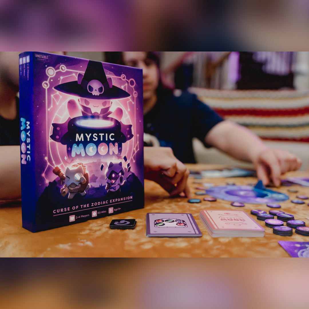 Mystic Moon Curse of the Zodiac Expansion - the box is purple and has a cat witch with a cauldron above two other cat characters. IN the background you can see people playing the game. 