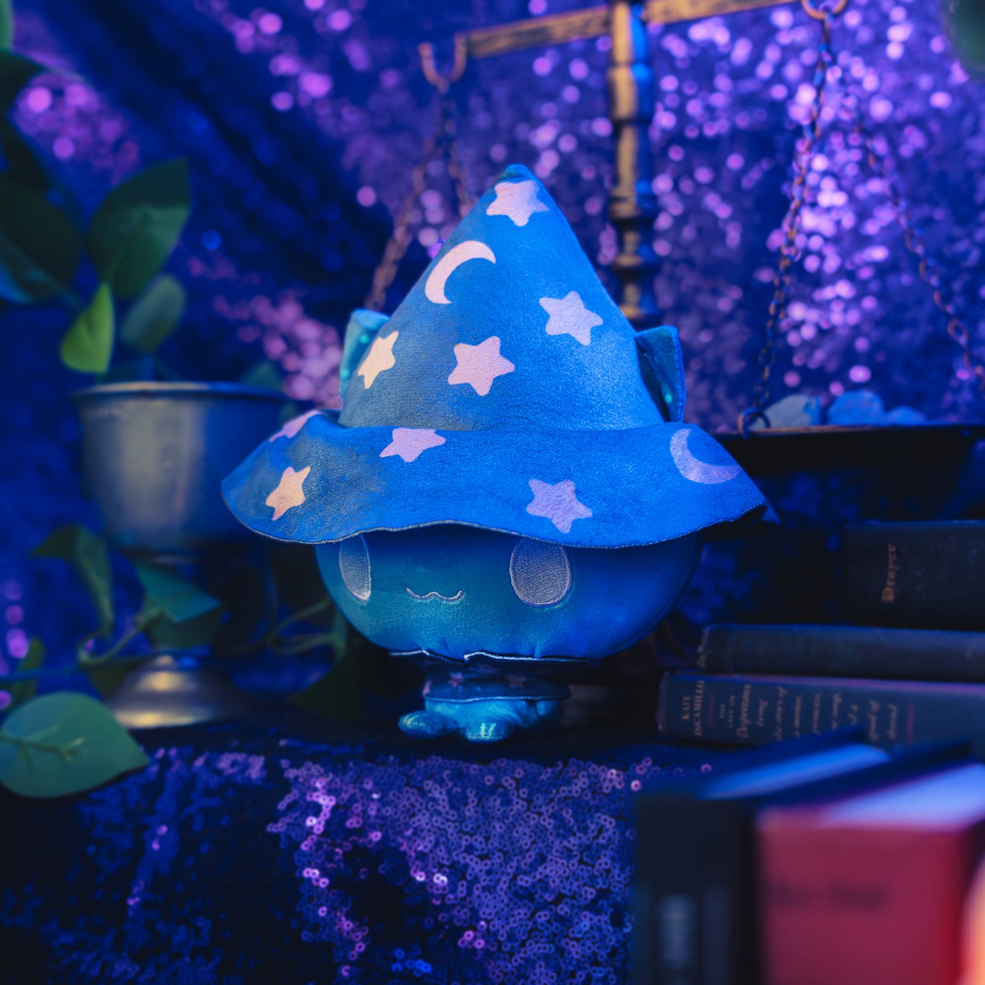 Cloudheads: 5" Mystic Moon (Eclipse) -A blue plush cat wearing a dark blue witch hat with tan stars and crescent moon patterns, shown with a sparkly purple backdrop as well as a leafy green plant and a pile of books. 
