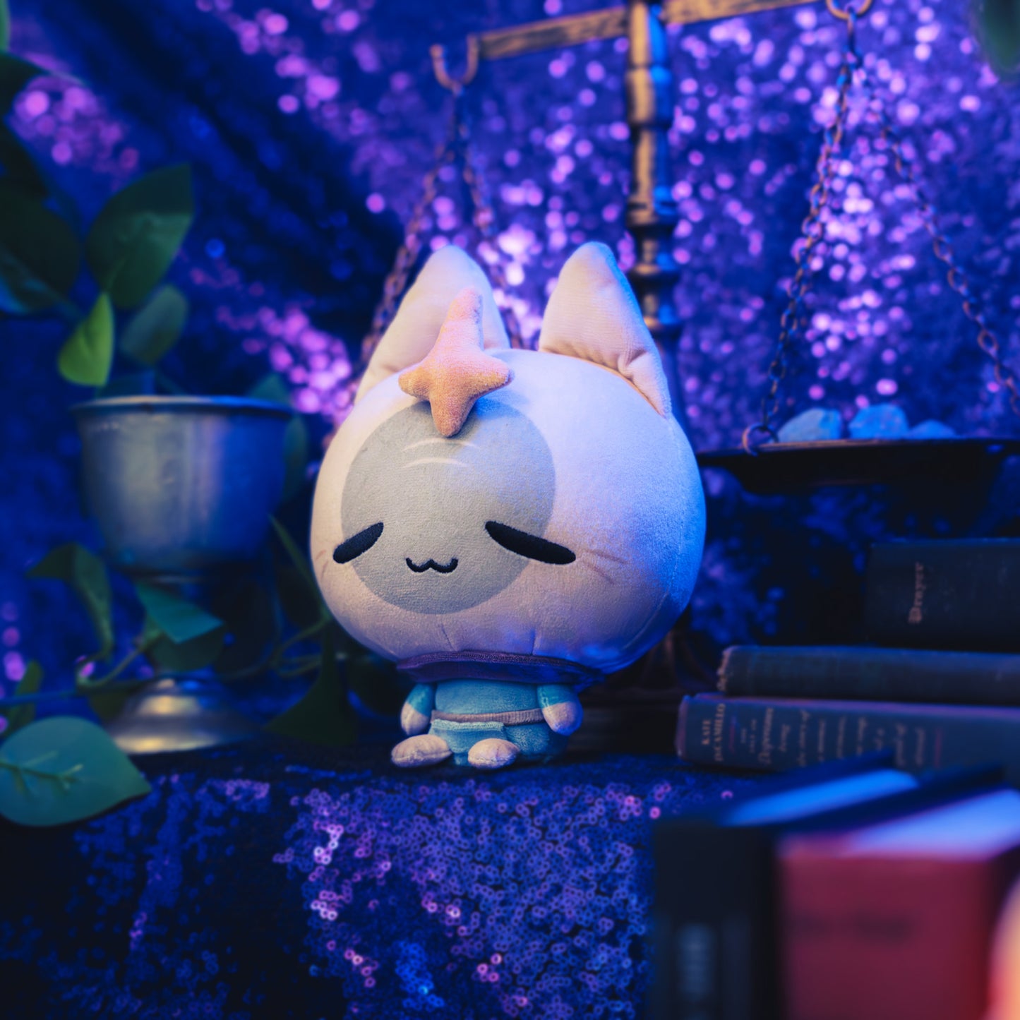 Cloudheads: 5" Mystic Moon (Elder Witch) -  A plush cat with a beige head, gray face, orange star on its forehead, and a blue outfit, shown with a sparkly purple backdrop as well as a leafy green plant and a pile of books. 
