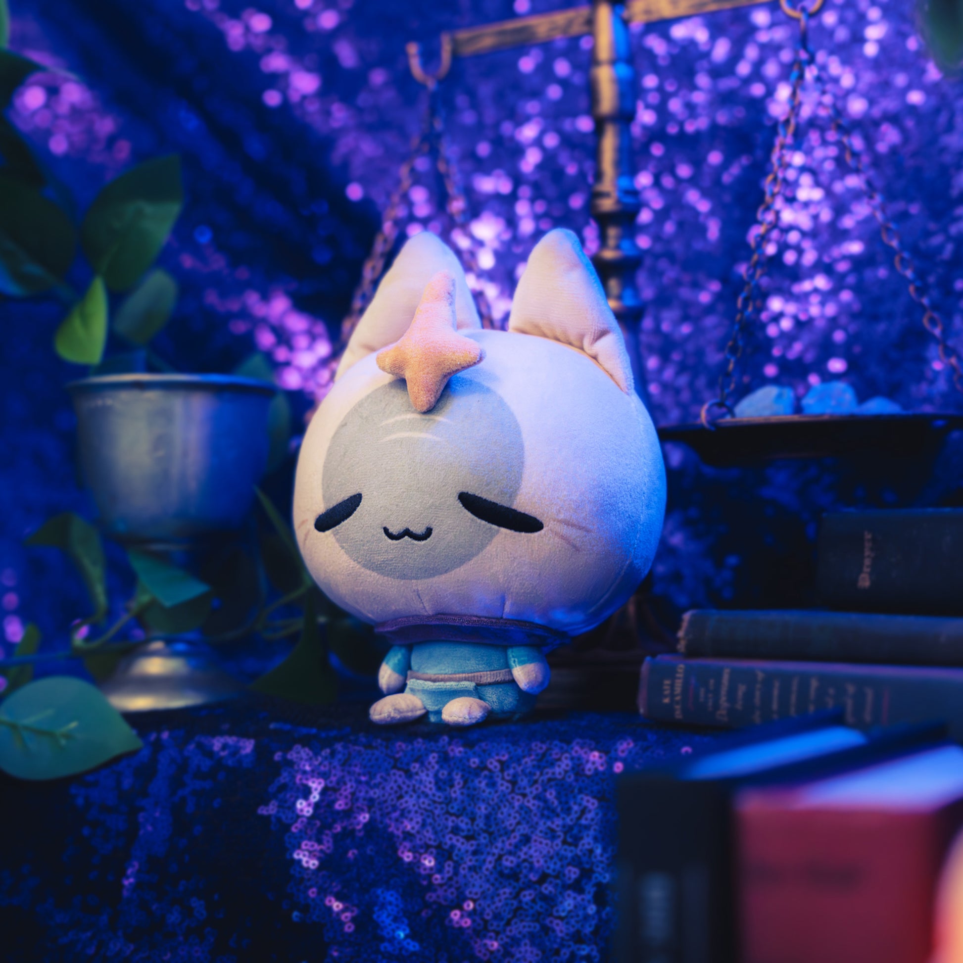 Cloudheads: 5" Mystic Moon (Elder Witch) -  A plush cat with a beige head, gray face, orange star on its forehead, and a blue outfit, shown with a sparkly purple backdrop as well as a leafy green plant and a pile of books. 
