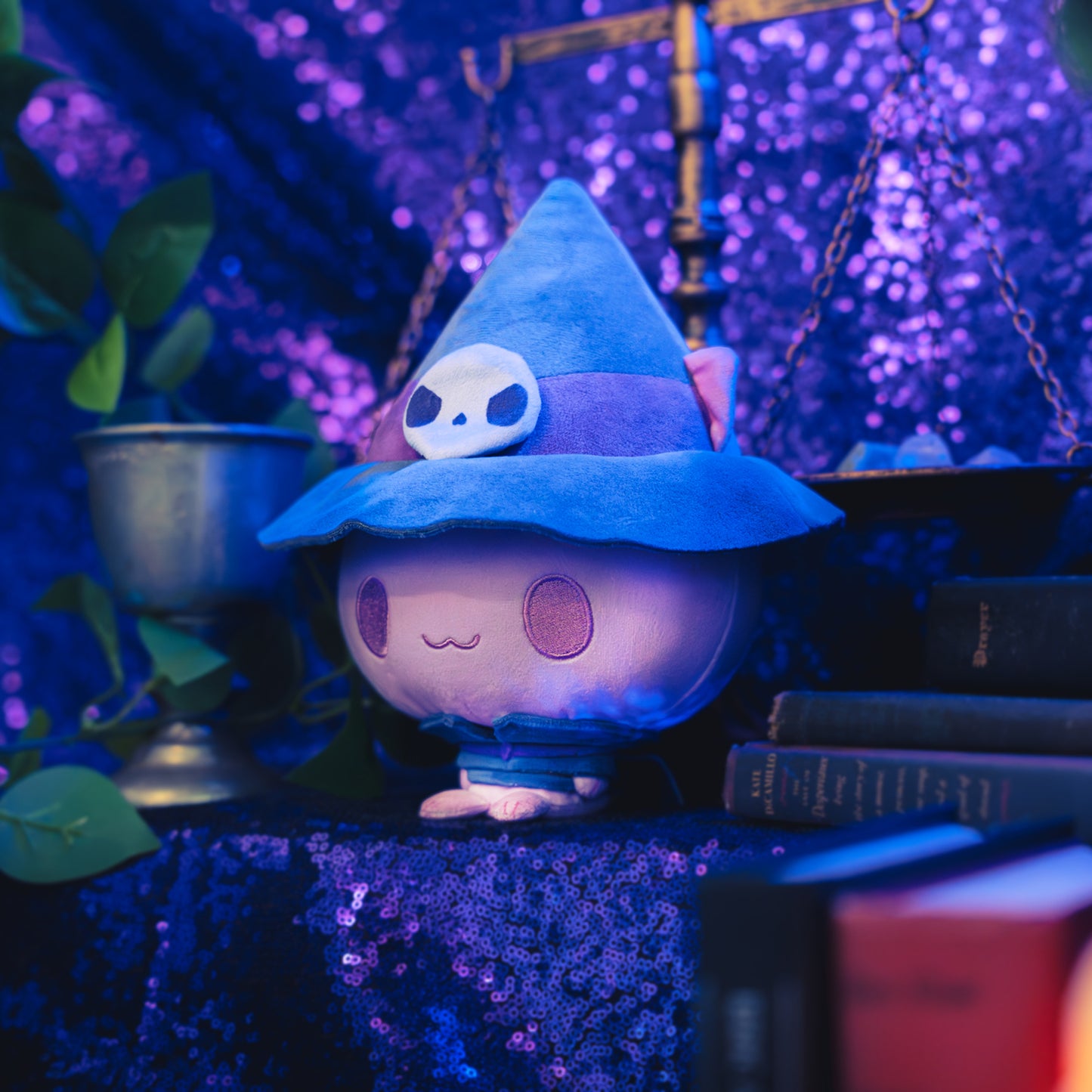 Cloudheads: 5" Mystic Moon (Jinx) - A pink plush cat with a witch hat, featuring a purple band with a white skull and a small purple sash, shown with a sparkly purple backdrop as well as a leafy green plant and a pile of books. 
