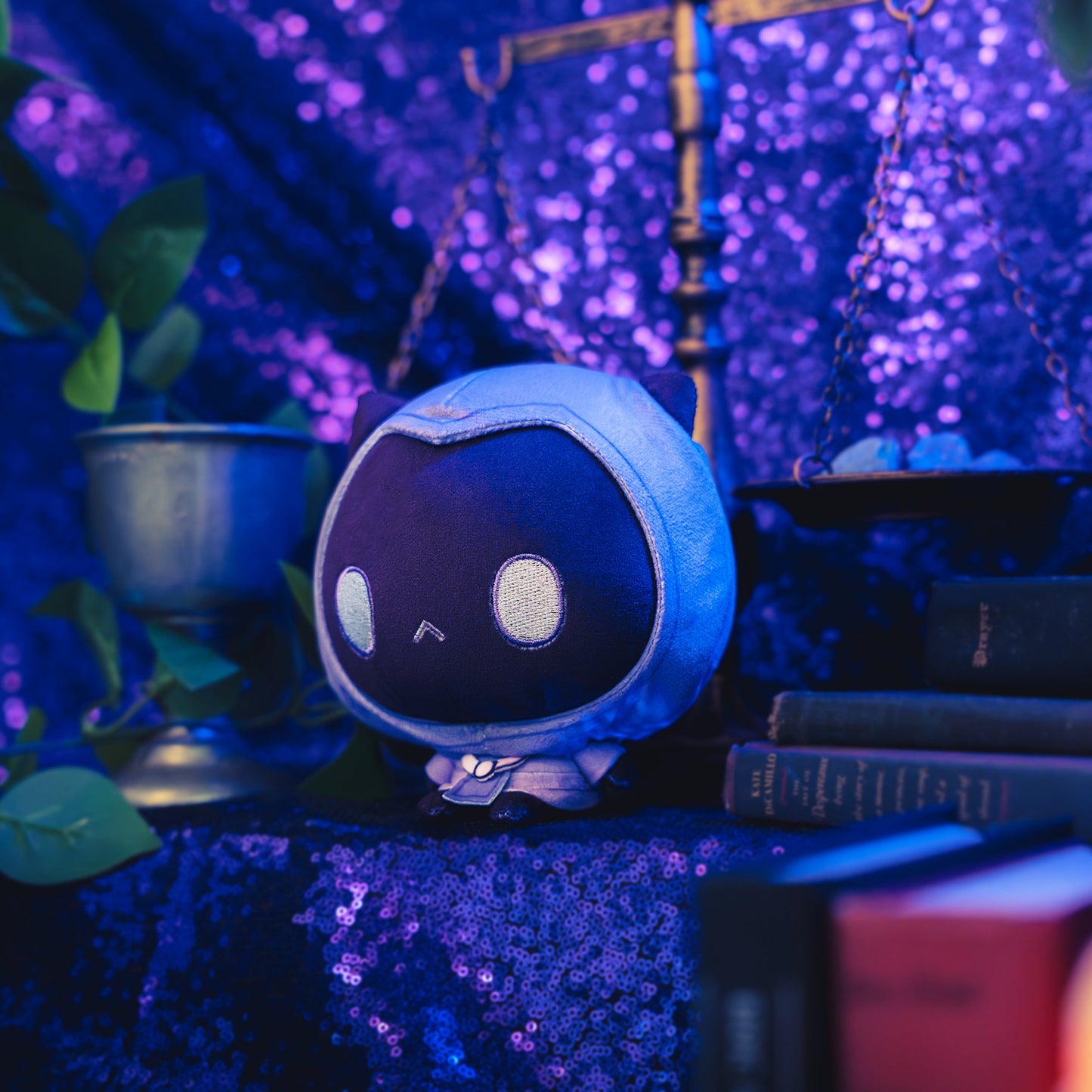Cloudheads: 5" Mystic Moon (Phantom). A purple and dark purple plush cat with large white oval eyes, and a small stitched frown, shown with a sparkly purple backdrop as well as a leafy green plant and a pile of books. 