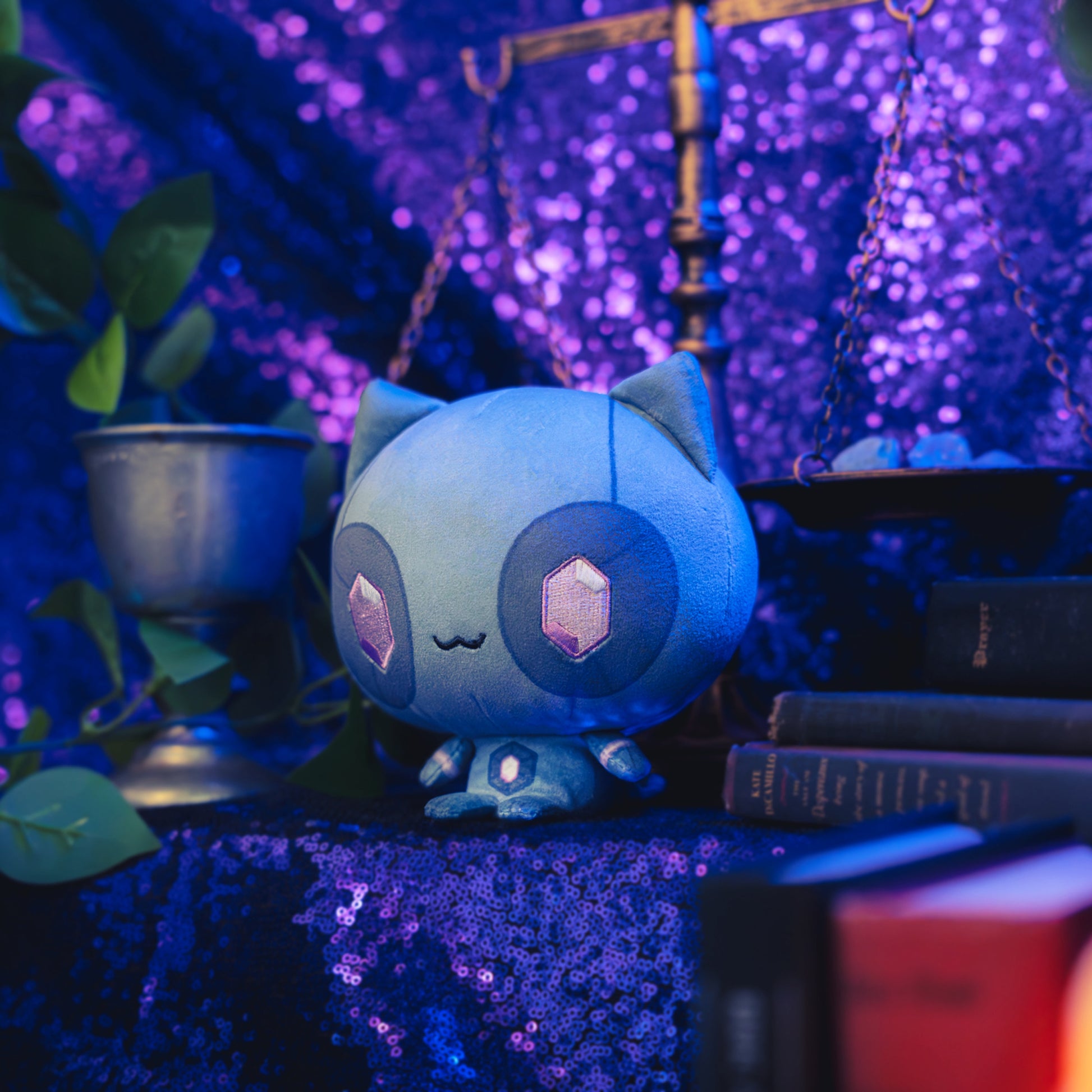 Cloudheads: 5" Mystic Moon (Quartz) - A blue cat plush toy with large dark blue eyes with pink gem-shaped crystals in the center, and a small black stitched smile, shown with a sparkly purple backdrop as well as a leafy green plant, a goblet and a pile of books. 