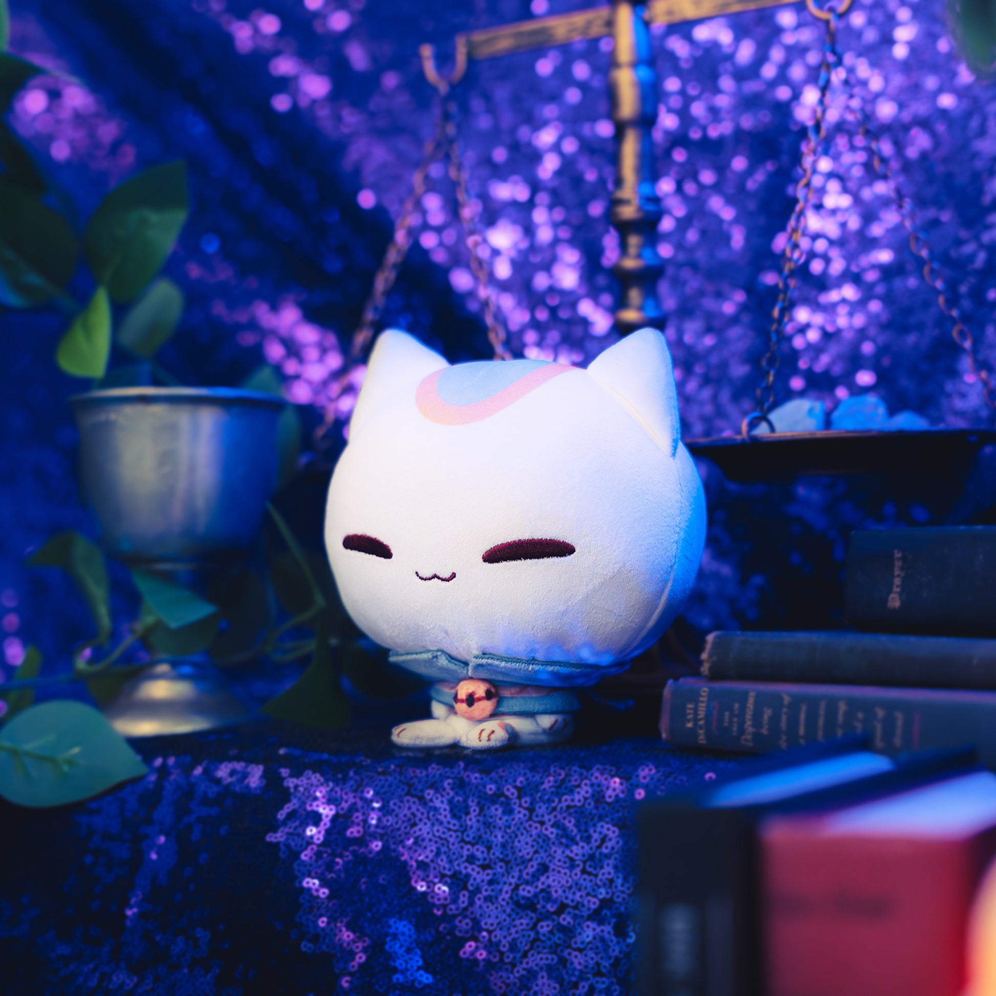 Cloudheads: 5" Mystic Moon (Vampurr). A white plush cat with closed eyes, a blue-gray collar, an orange accent on its head, and a smirk, shown with a sparkly purple backdrop as well as a leafy green plant and a pile of books. 
