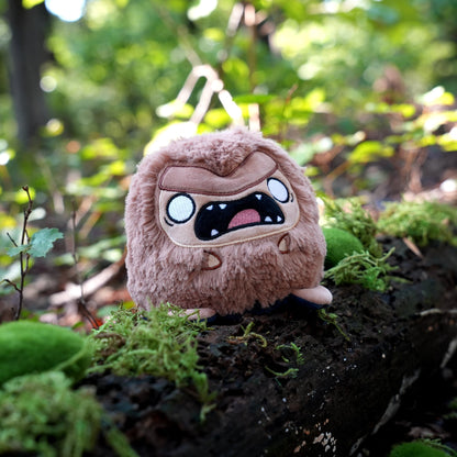 Twisted Cryptids: Bigfoot 4" Reversible Plushie angry side sitting on a mossy log in a forest