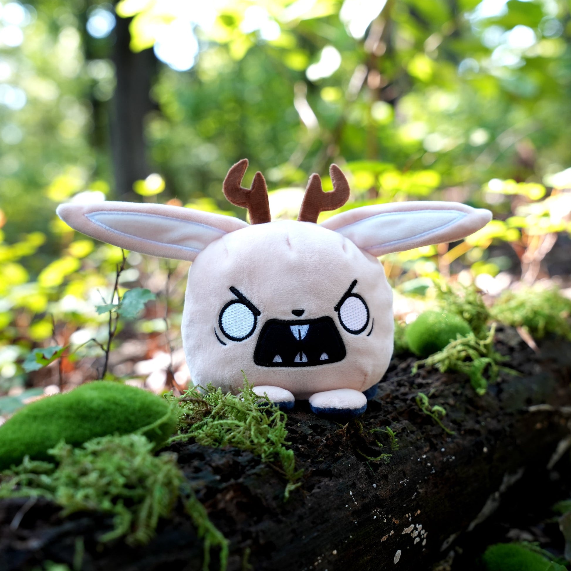 Twisted Cryptids: Jackalope 4" Reversible Plushie angry side sitting on a mossy log in a forest