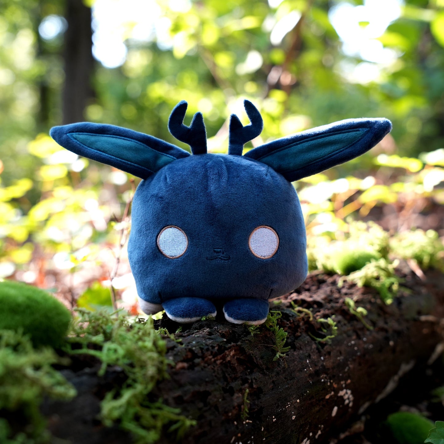 Twisted Cryptids: Jackalope 4" Reversible Plushie happy side sitting on a mossy log in a forest