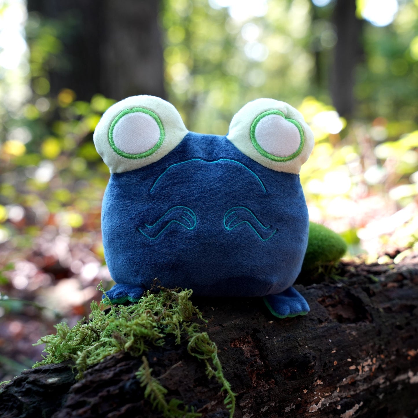 Twisted Cryptids: Loveland Frog 4" Reversible Plushie sad side sitting on a mossy log in a forest