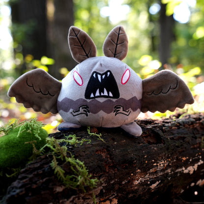 Twisted Cryptids: Mothman 4" Reversible Plushie angry side sitting on a mossy log in a forest
