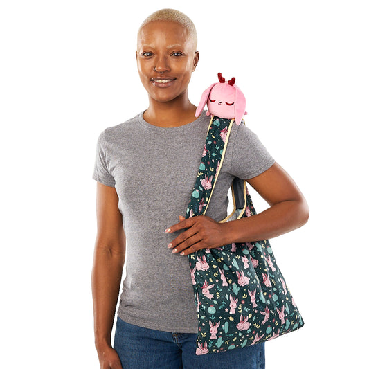 Woman with a close-cropped hairstyle, smiling and wearing a gray t-shirt, carrying a TeeTurtle Plushiverse Fearsome Jackalope Plushie Tote Bag with a pink TeeTurtle plushie.