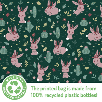 Patterned fabric design featuring pink rabbits, green cacti, and small floral elements on a dark green background, noted as made from 100% recycled plastic bottles. Part of the TeeTurtle Plushiverse Fearsome Jackalope Plushie Tote Bag.