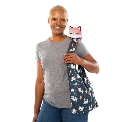 A smiling bald woman in a gray t-shirt and jeans, carrying a navy Plushiverse Enchanting Kitsune Tote Bag with cat designs, with a TeeTurtle plushie peeking out.