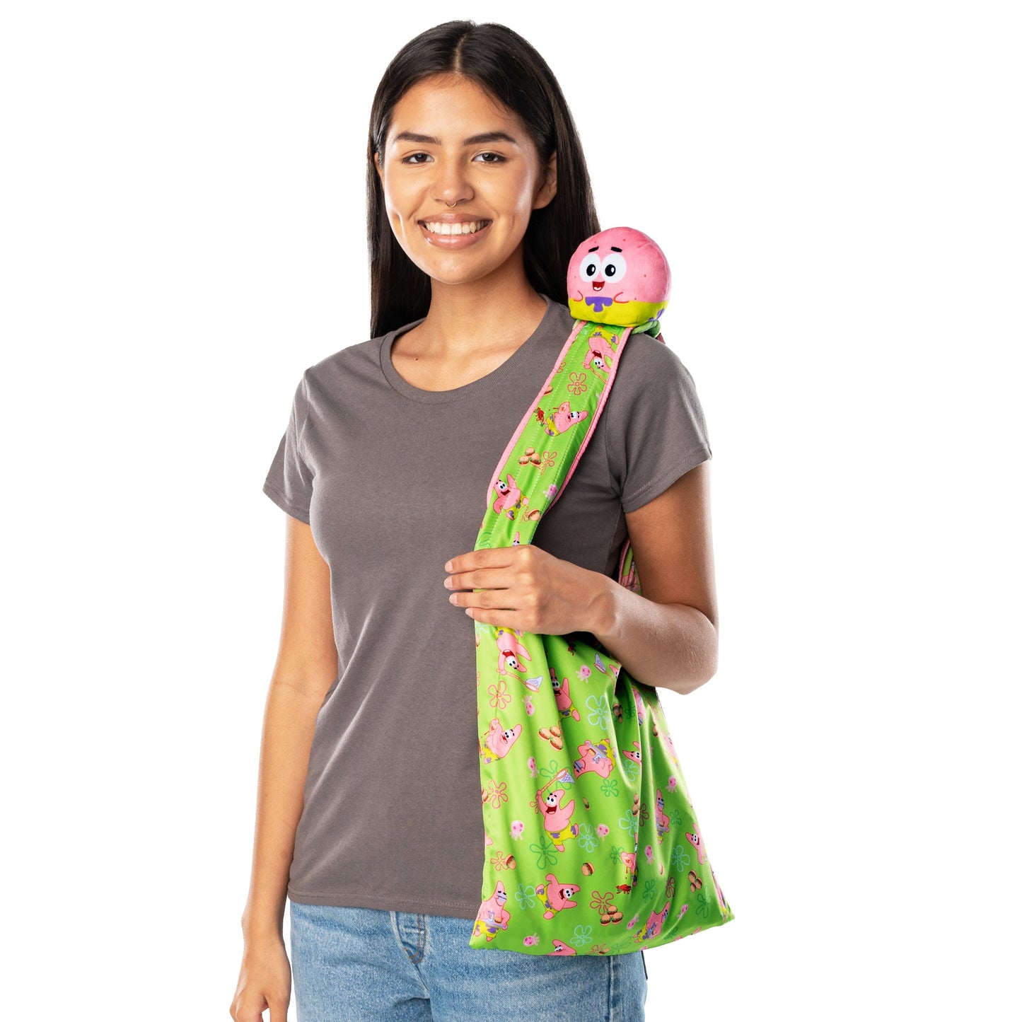 A woman in a gray shirt and jeans holds a Plushiverse Patrick Star Snacktime Plushie Tote Bag crafted from recycled plastic bottles, adorned with a round pink Nickelodeon plushie, smiling at the camera.